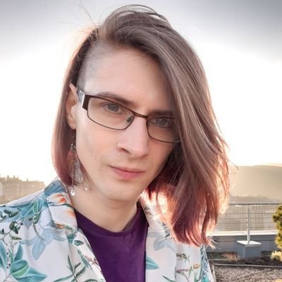 A headshot of Sy standing on top of a building. Sy has long brown hair and is wearing a white flowery shirt over a purple t-shirt, and a feather earing